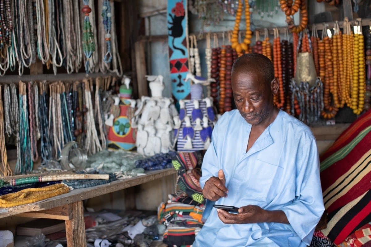 A Nigerian market trader checking his phone at his stall, surrounded by beads and crafts